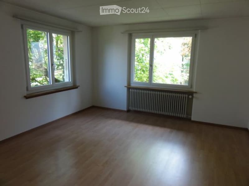 Empty room with wooden flooring, white walls, and two windows with wooden sills. A radiator is visible under one window.
