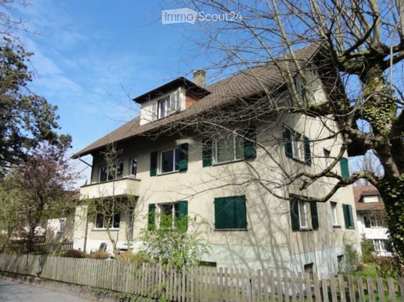 A two-story residential house with a pitched roof, green shutters, and a wooden fence in front.