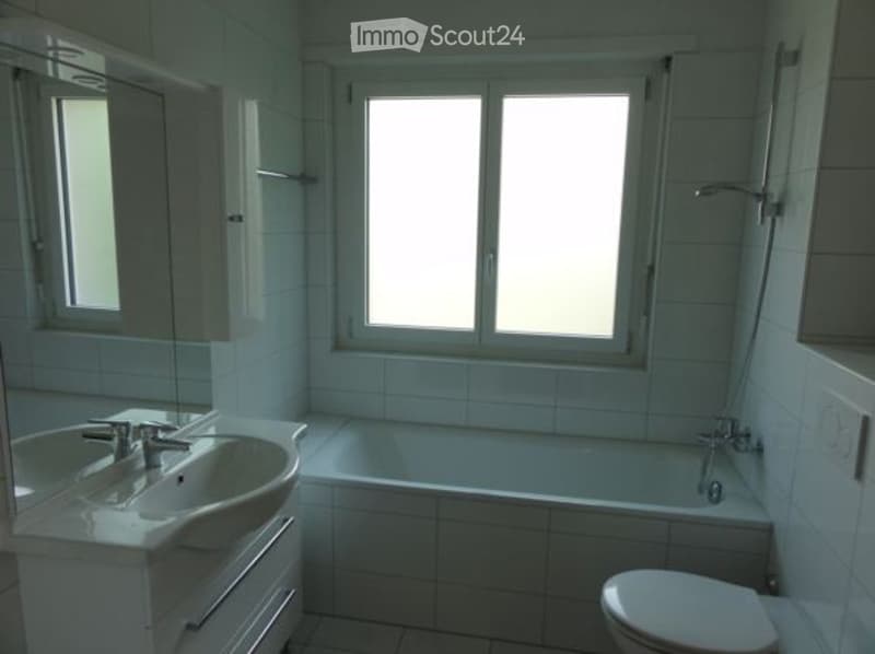 Modern bathroom with white tiles, a sink with a faucet, a bathtub, and a toilet. The room has large windows providing natural light.
