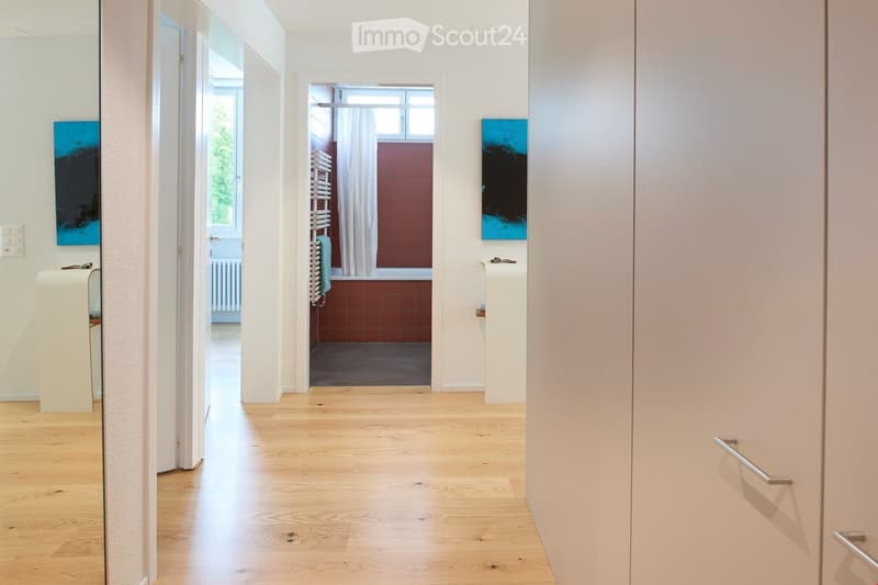 Bathroom with shower, towel radiator, toilet paper holder, blue artworks, wooden floor, and mirror in hallway.