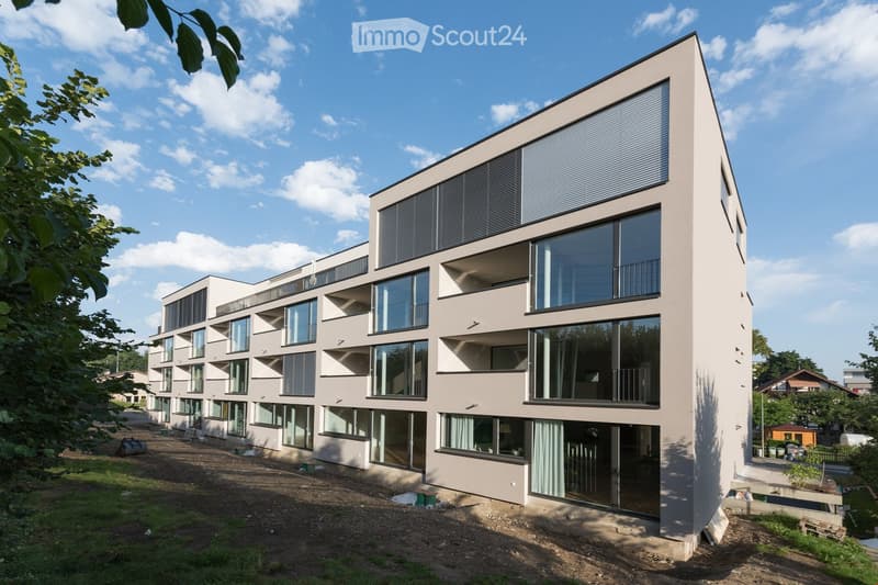 Modern apartment building, large glass windows, balconies, raised ground floor, beige exterior