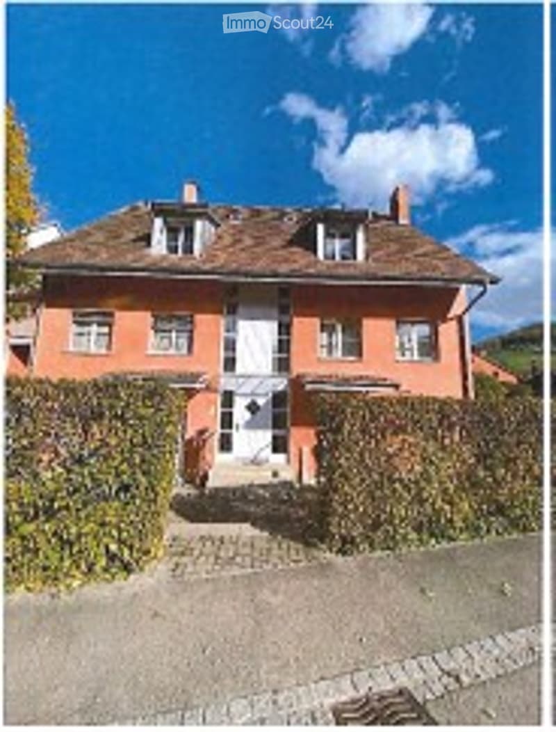 Two-story house with red brick facade, white-framed windows, and a central entrance door. The roof is covered with brown tiles and has multiple chimneys. The house is surrounded by a neatly trimmed hedge and is set against a backdrop of a clear blue sky w