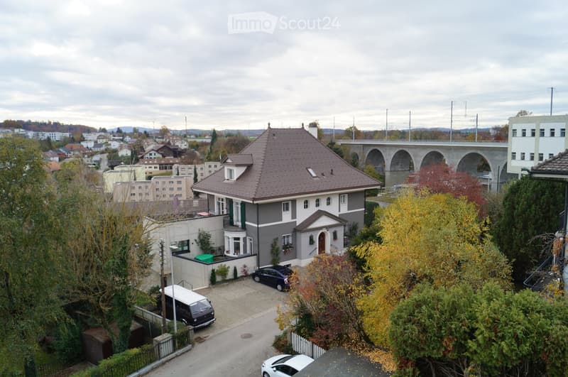 A two-story house with a brown tiled roof, surrounded by trees and parked cars, with a bridge visible in the background.