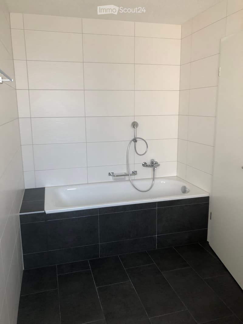 Modern bathroom with a white bathtub, black tiled floor, and white tiled walls. Features a showerhead and faucet above the tub.