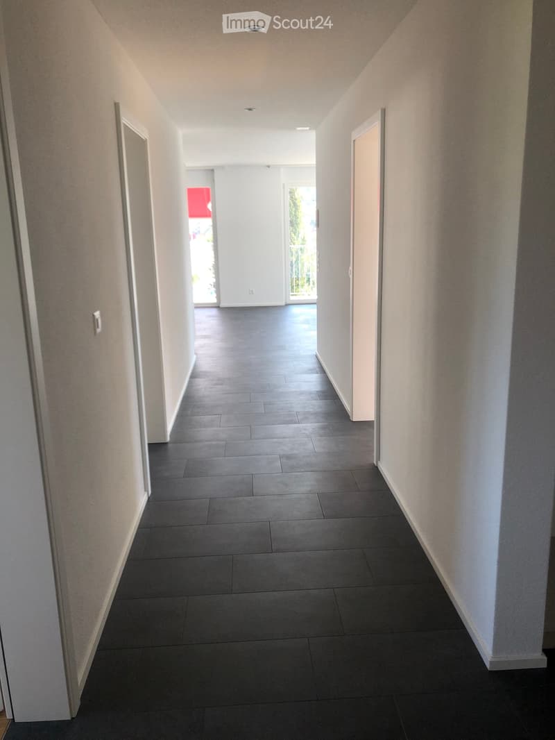 Empty hallway with grey tiled floor, white walls, and recessed lighting. Doors lead to other rooms and an outdoor area.