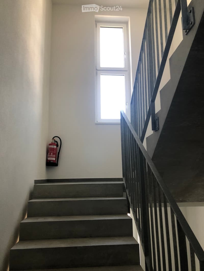 Staircase with grey concrete steps, metal railing, and a window at the top. A red fire extinguisher is mounted on the wall to the left.