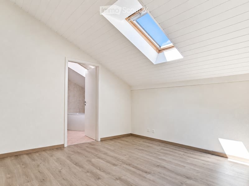 Empty attic room with wooden flooring, white walls, and a skylight window. An open doorway leads to a small bathroom with a bathtub.