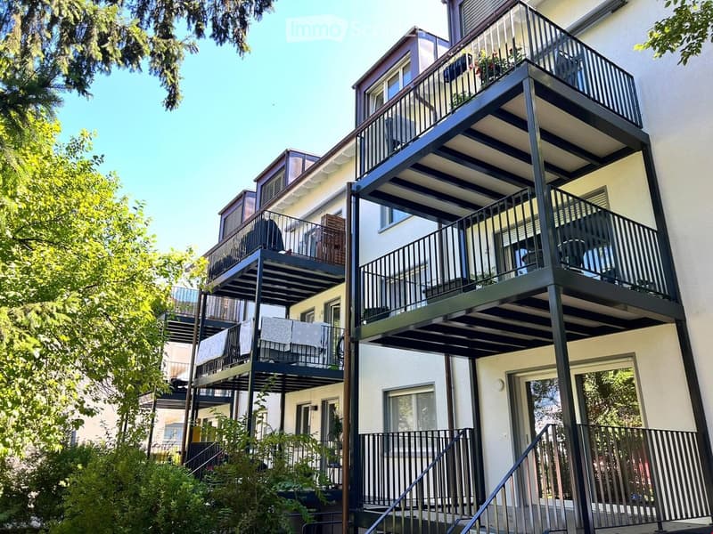 Multi-story residential building with multiple balconies, modern railings, and greenery around the base.