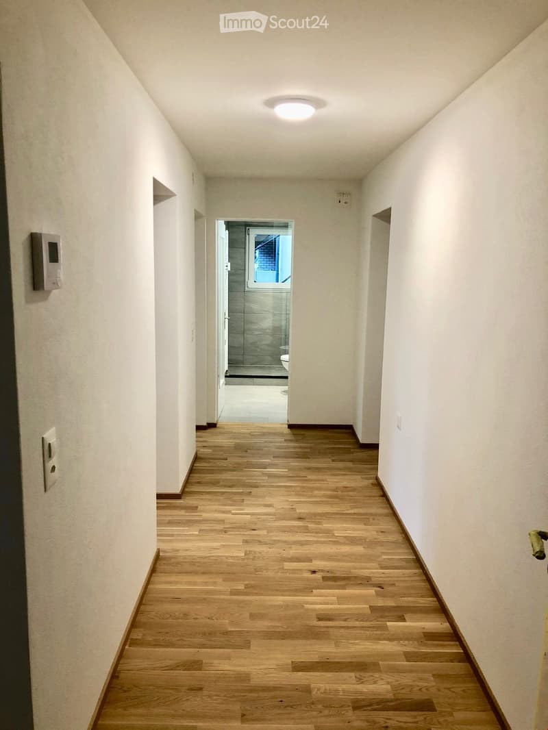 Clean, well-lit hallway with wooden flooring, white walls, and a ceiling light fixture. An open doorway at the end leads to a tiled bathroom area.