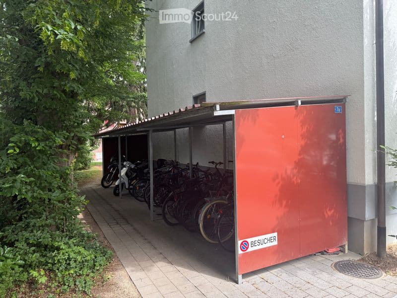 A covered parking area with several bicycles and a scooter parked inside, adjacent to a building with a sign indicating visitor access.