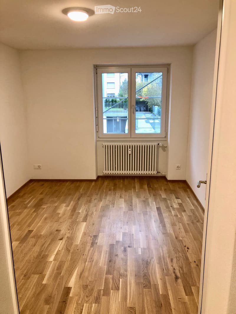 Empty room with light wooden flooring, white walls, a radiator, and a large window with a view of a balcony and outdoor area.