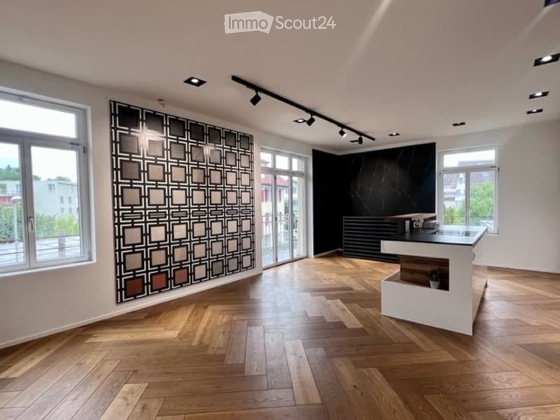 Modern reception area with herringbone wooden flooring, white reception desk, decorative wall art, and large windows offering a view of the outside.