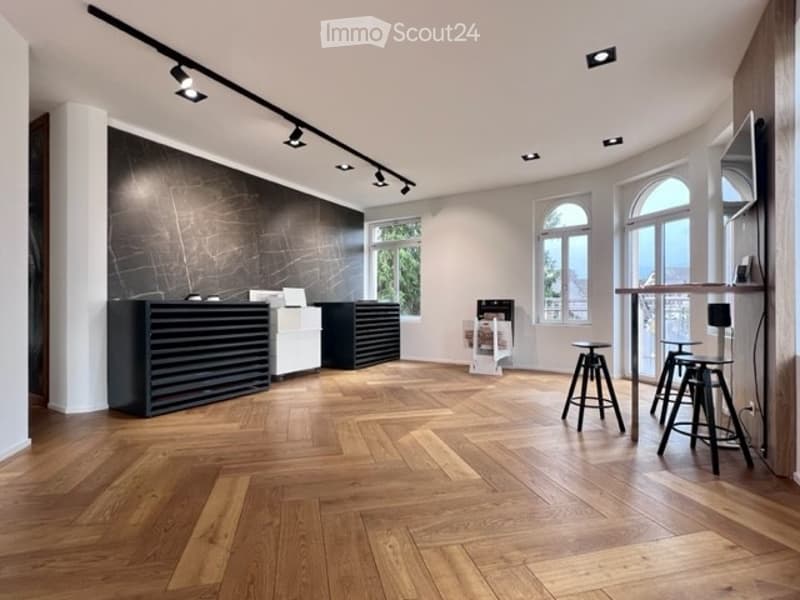 Modern, minimalist room with a reception desk, two stools, a table, and a screen. The room features large arched windows, wooden flooring, and track lighting.