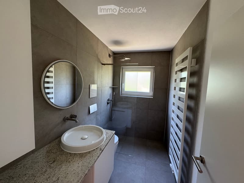 Modern bathroom with a white sink, round mirror, glass shower enclosure, and a radiator. The space features grey tiles and a window providing natural light.
