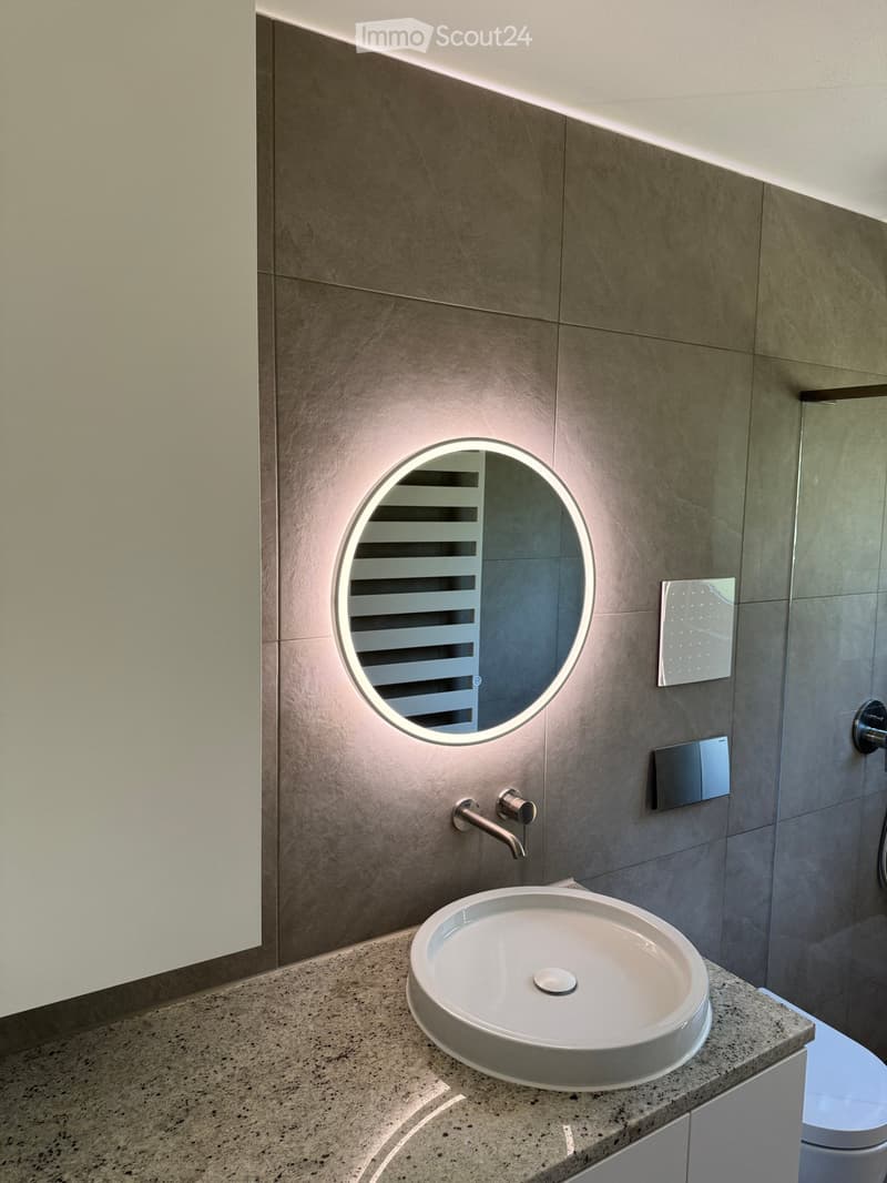 Modern bathroom with a round sink, sleek faucet, and illuminated round mirror above it. Grey tiled walls and a granite countertop.