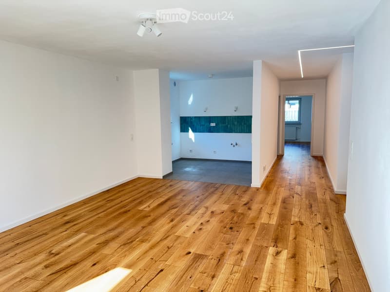Empty hallway with wooden flooring, white walls, and recessed ceiling lighting. The hallway leads to a tiled room and a windowed area with a radiator.