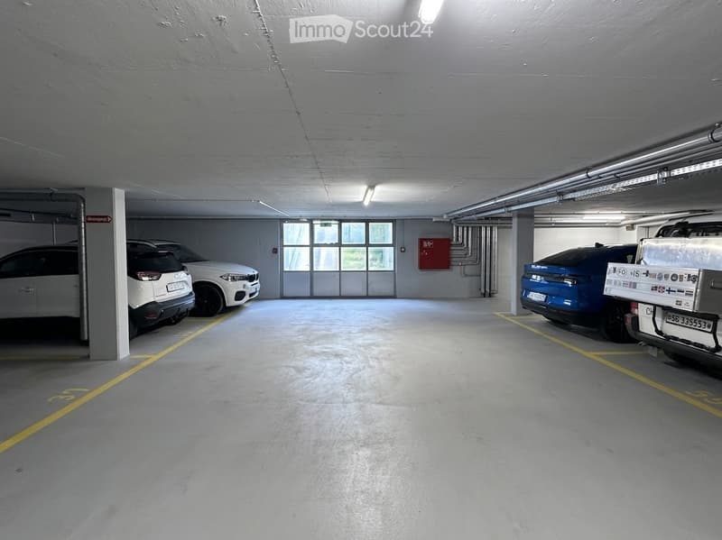 Underground parking area with several parked cars, marked parking spaces, and fluorescent lighting.