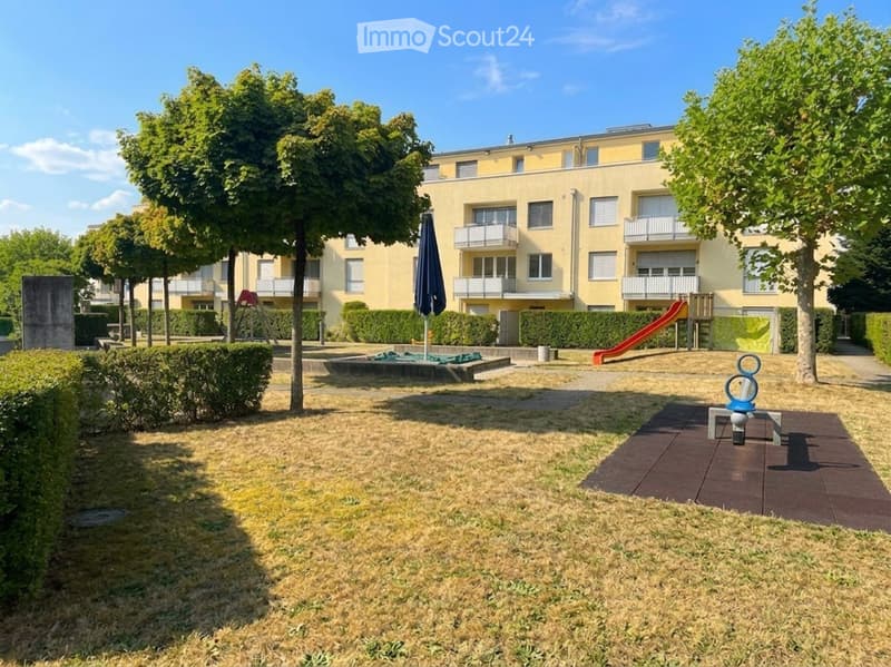 Courtyard area with a small fountain, playground equipment, and a multi-story residential building in the background.