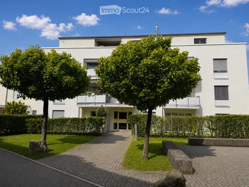 Modern two-story white building with balconies, surrounded by greenery and a paved walkway.
