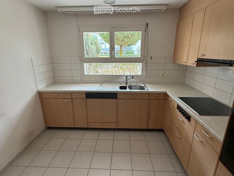 Modern kitchen with light wooden cabinets, tiled floor, and a window overlooking greenery.