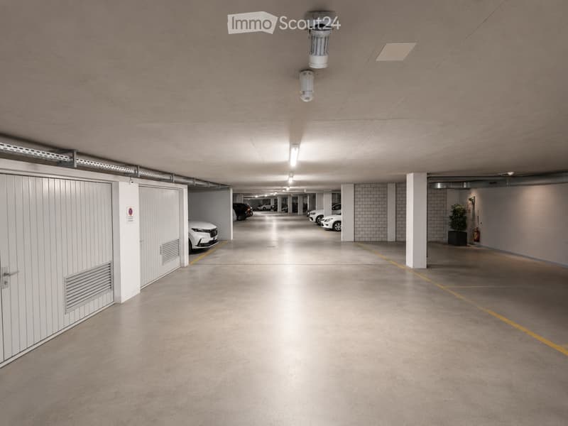 Underground parking area with several parked cars, concrete floors, white pillars, and overhead lighting.