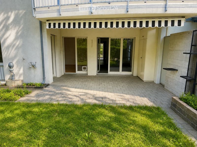 Brick-paved outdoor area with a grassy lawn, small plants, and a covered entrance to a building with large glass doors and a striped awning.