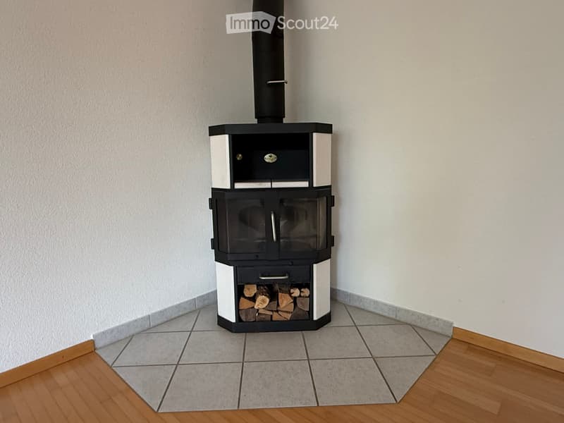 Corner fireplace with wood storage compartment and chimney, placed on tiled floor adjacent to wooden flooring.