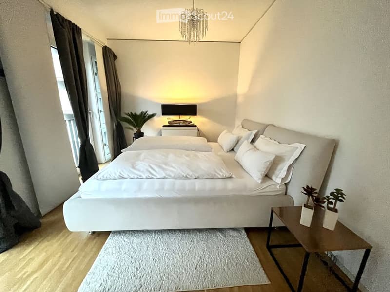 Modern bedroom with a large bed, nightstand, table lamp, side table with potted plants, and a view of a balcony.