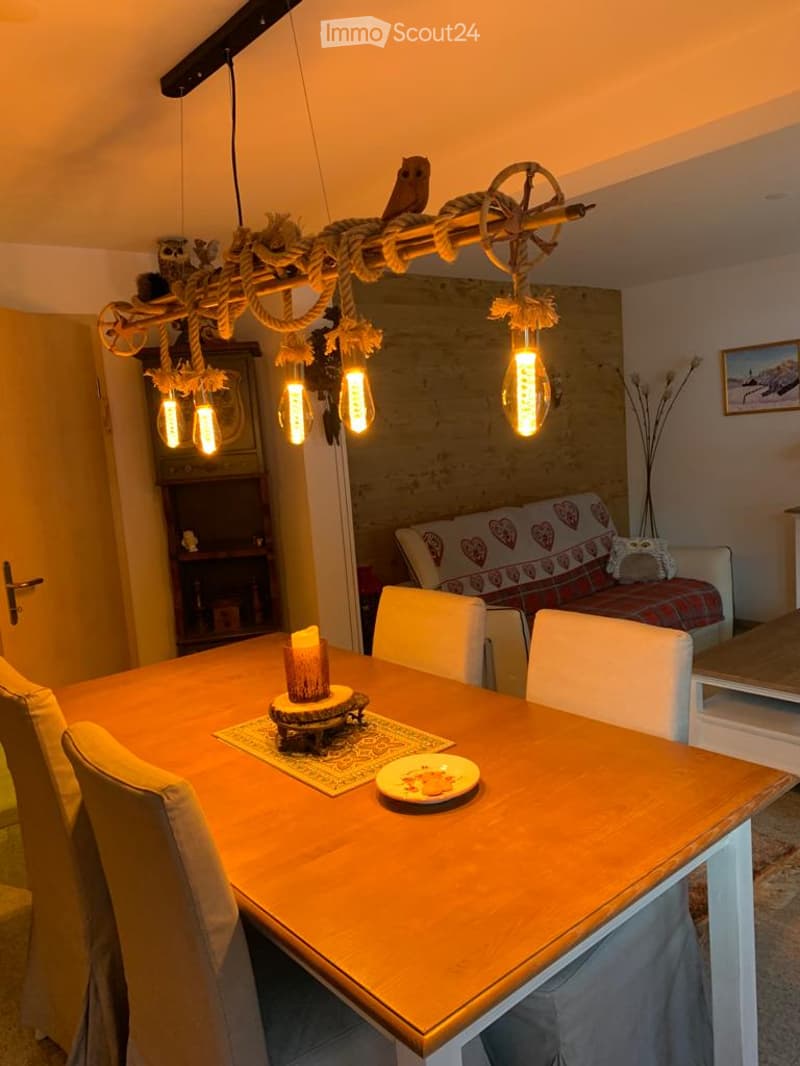 A cozy dining and living area with a wooden dining table, chairs, a sofa with a patterned throw, a cabinet, and a unique hanging light fixture with exposed bulbs and rope details.