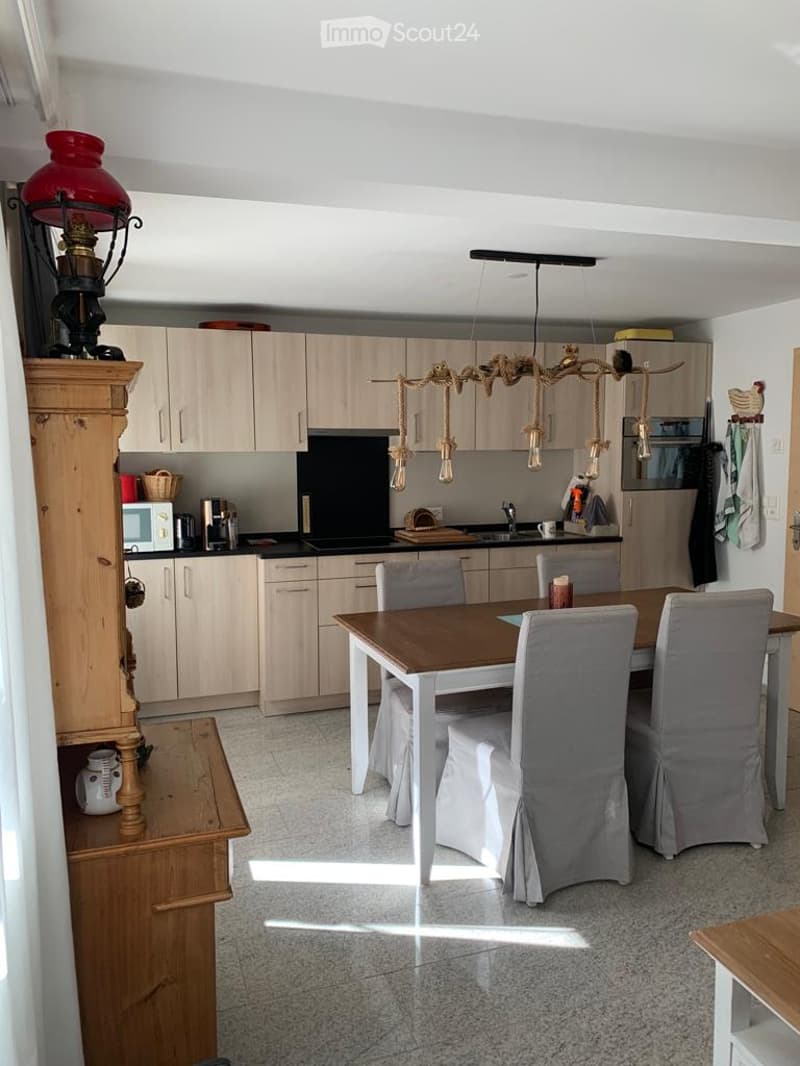 Bright kitchen with light wood cabinets, a central dining table with chairs, a microwave, and a hanging light fixture.