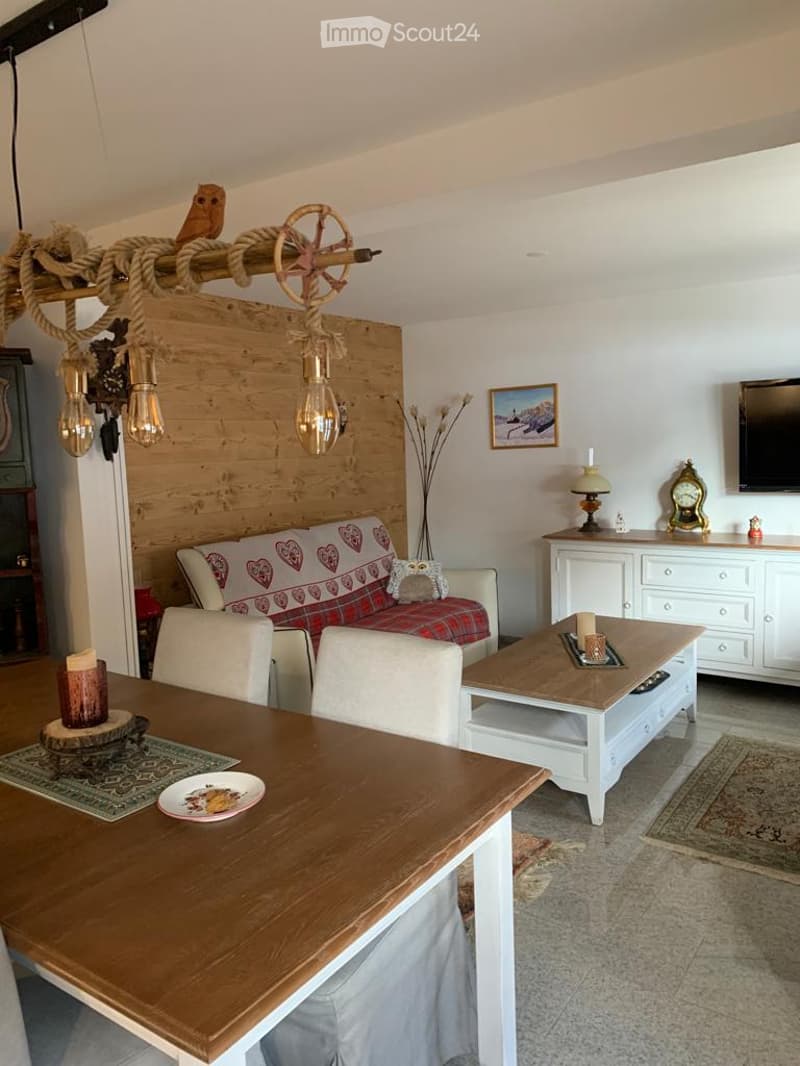 Cozy living room with a red and white patterned sofa, wooden coffee table, dining table with chairs, and a television mounted on the wall. The room features a wooden accent wall, a decorative chandelier, and a small side table with a lamp.