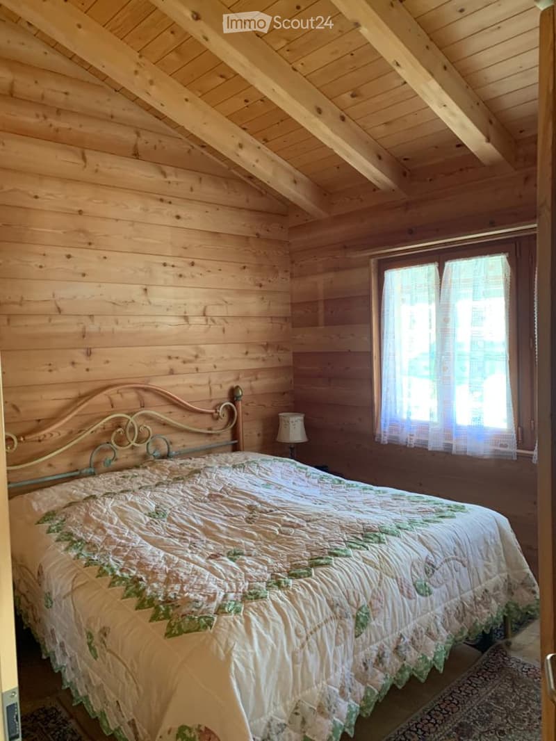 Cozy wooden bedroom with exposed beams, a single bed with a floral quilt, a small table lamp, and a window with sheer curtains.