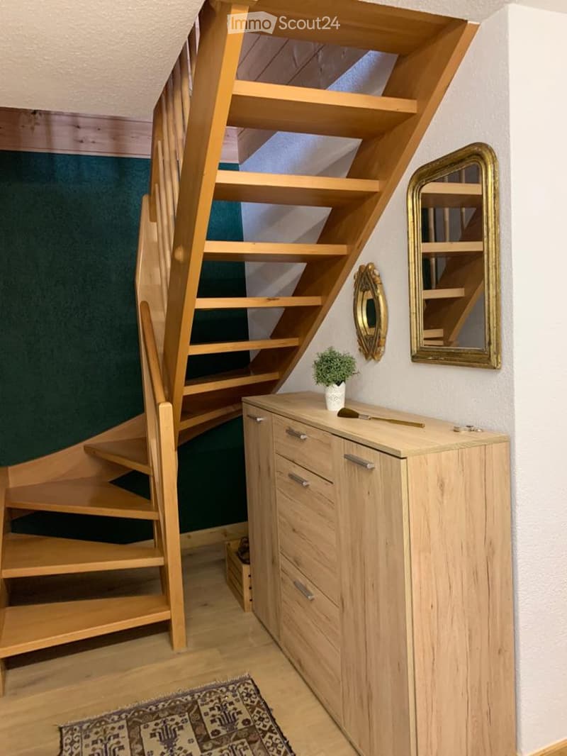 A compact, well-lit hallway features a wooden staircase with built-in shelves, a light wooden cabinet with drawers, a small potted plant, and decorative mirrors on the wall.