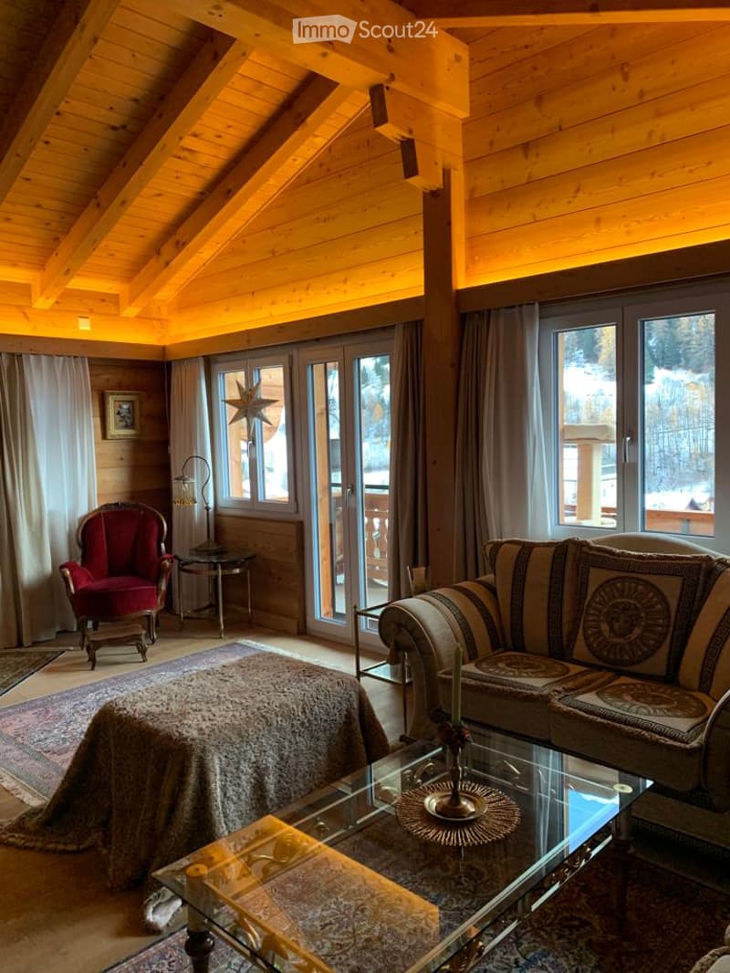 Cozy living room with wooden beams, large windows, a sofa, armchair, coffee table, and a view of the snowy landscape.