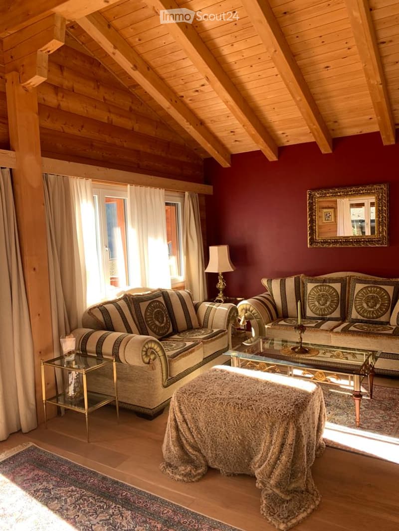 Cozy living room with exposed wooden beams, dark red walls, large windows with white curtains, patterned sofa, glass coffee table, floor lamp, and decorative mirror.
