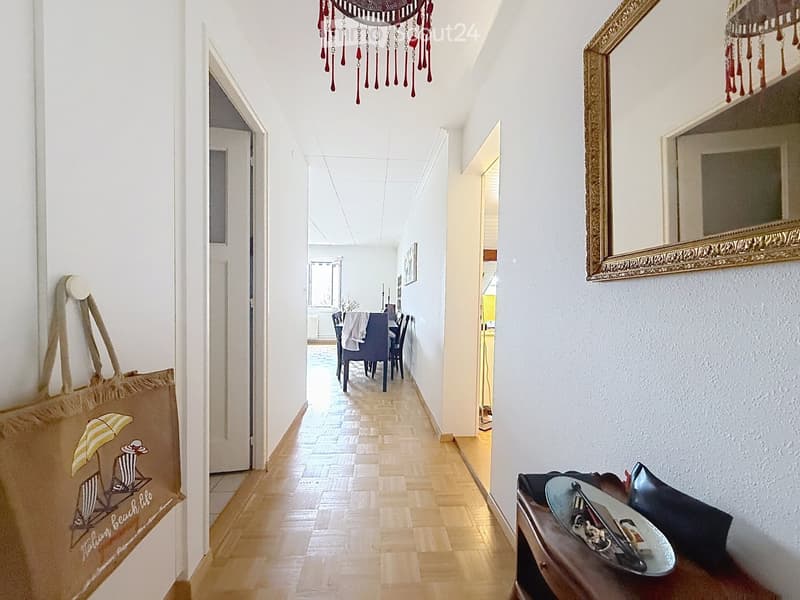 A well-lit hallway with wooden flooring, white walls, and a decorative hanging ornament. A large mirror with an ornate frame is mounted on the wall, and a woven beach-themed bag hangs on the left side. A small bench with a folded jacket and a pair of skis