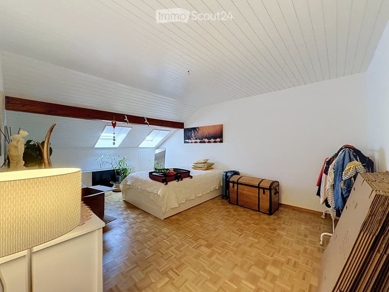 Bright bedroom with wooden flooring, slanted white ceiling, skylight windows, bed with white linens, decorative trunk, clothing rack, and a plant near the window.