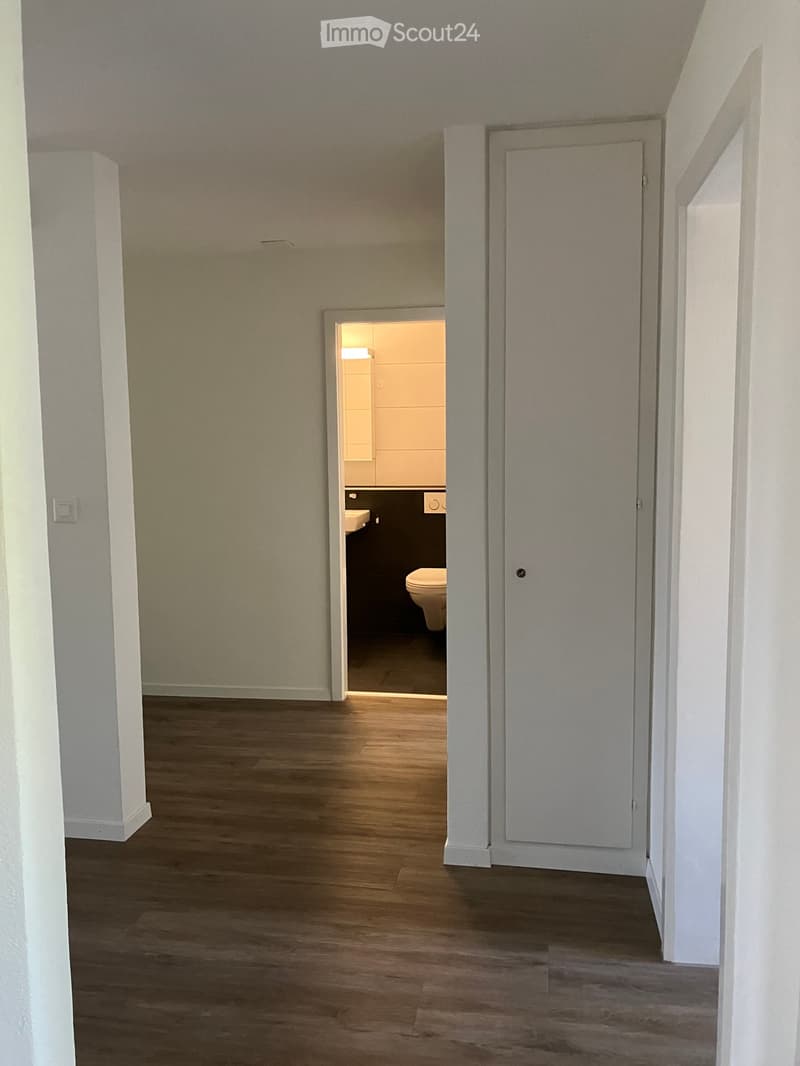 Empty hallway with wooden flooring, white walls, and a partially open door leading to a small bathroom with a toilet and sink.