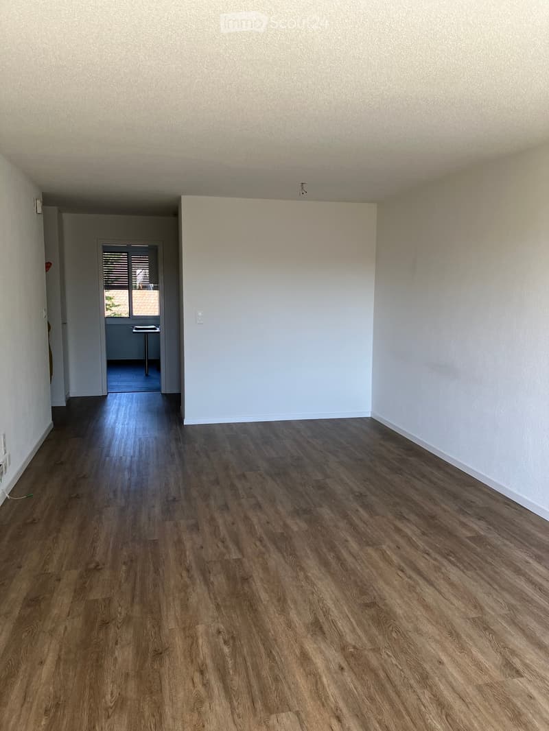 Empty room with wooden flooring and white walls, featuring a doorway leading to another room with a sliding glass door and a small table visible in the adjacent space.