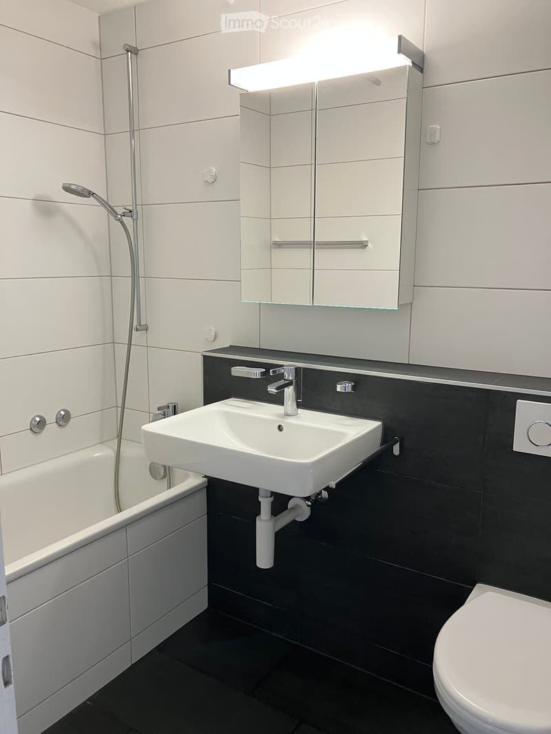 Modern bathroom with white tiles, black lower half walls, a bathtub, a sink with a modern faucet, a large mirror with built-in lighting, and a toilet.