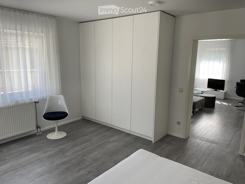Modern furnished room with a white wardrobe, a sleek white chair, a small table, and a TV screen in the adjacent area.