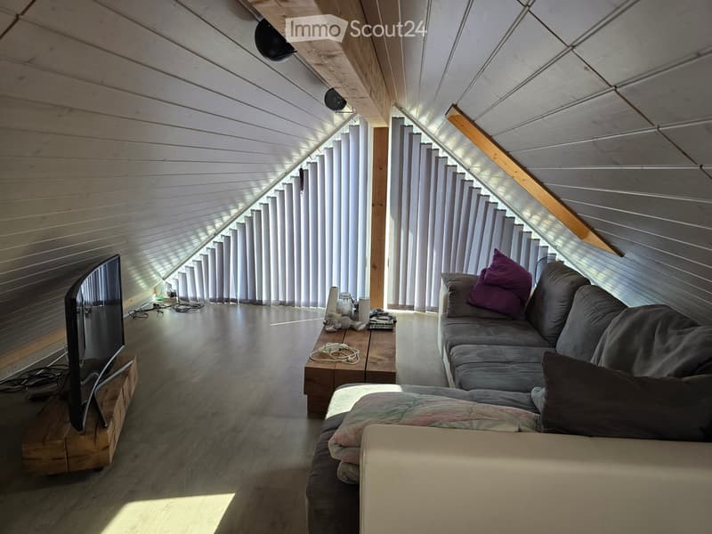 Living room with wooden floors, slanted wooden ceiling, large windows with blinds, sofa, coffee table, television, and table lamp.