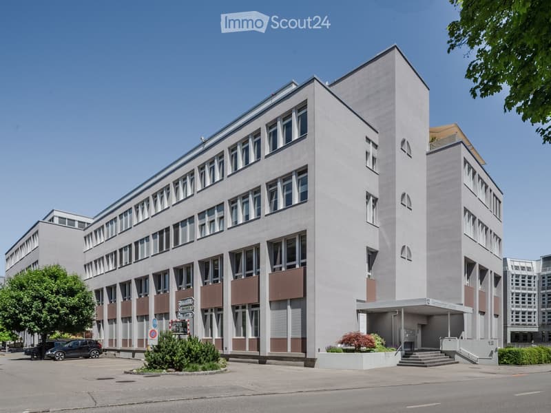 Modern multi-story building with light-colored facade, large windows, and geometric design. Features include a central entrance with steps, small landscaped area, and parked cars nearby.