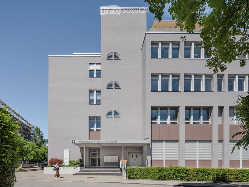 Modern multi-story building with light gray facade, large windows, and a central entrance labeled "Aerztzentrum Liebefeld".