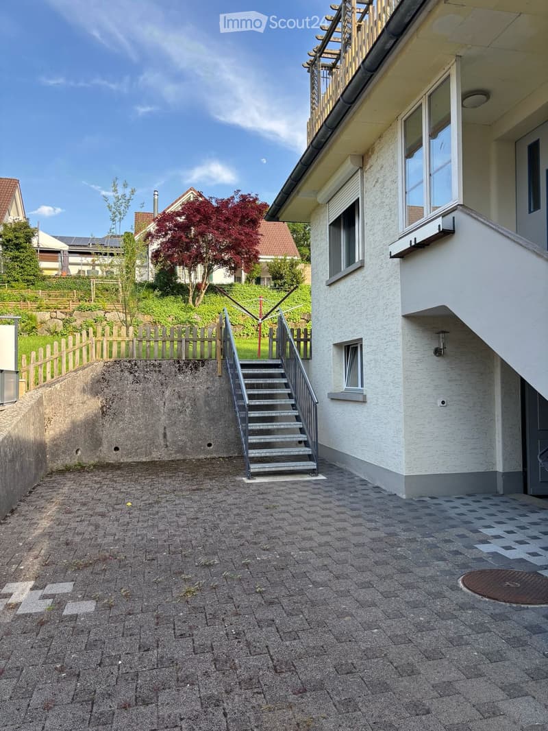 Paved outdoor area with steps leading up to a house, surrounded by greenery and a wooden fence.