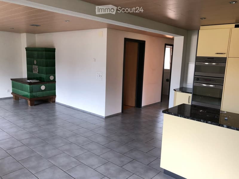 Modern kitchen with grey tiled floor, white walls, cream-colored cabinets, and a built-in oven. A green tiled stove sits on a wooden base to the left.