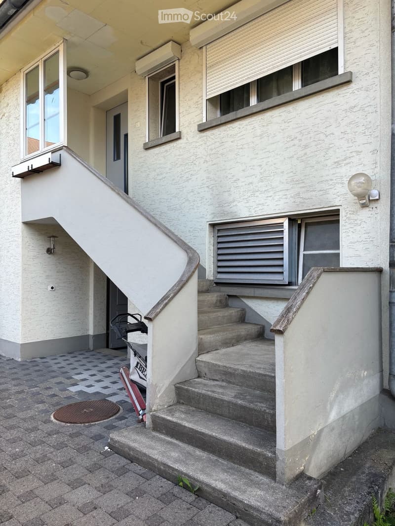 Exterior view of a raised ground floor entrance with concrete steps, handrails, and a small balcony area. The building has white walls, multiple windows, and a small staircase leading up to the entrance.