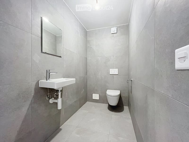 Modern bathroom with grey tiled walls and floor, white sink, chrome faucet, toilet, and a wall-mounted mirror.