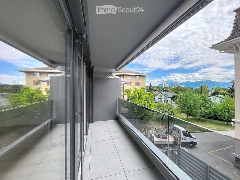 Modern balcony with large glass windows, tiled floor, and a view of a suburban area with trees, houses, and mountains.