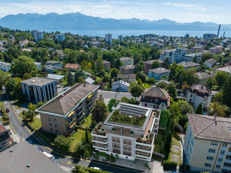 Aerial view of a residential area with multiple buildings, surrounded by greenery and a scenic lake and mountain backdrop.